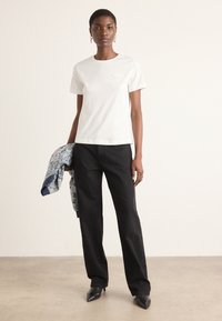 T-shirt bianco in cotone con piccolo dettaglio ricamato, abbinato a pantaloni neri a gamba dritta e una giacca con motivo blu drappeggiata sul braccio.