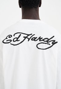 Weißes Langarmshirt mit großem, schwarzem besticktem "Ed Hardy"-Logo auf dem Rücken. Weicher Stoff mit lockerer Passform.
