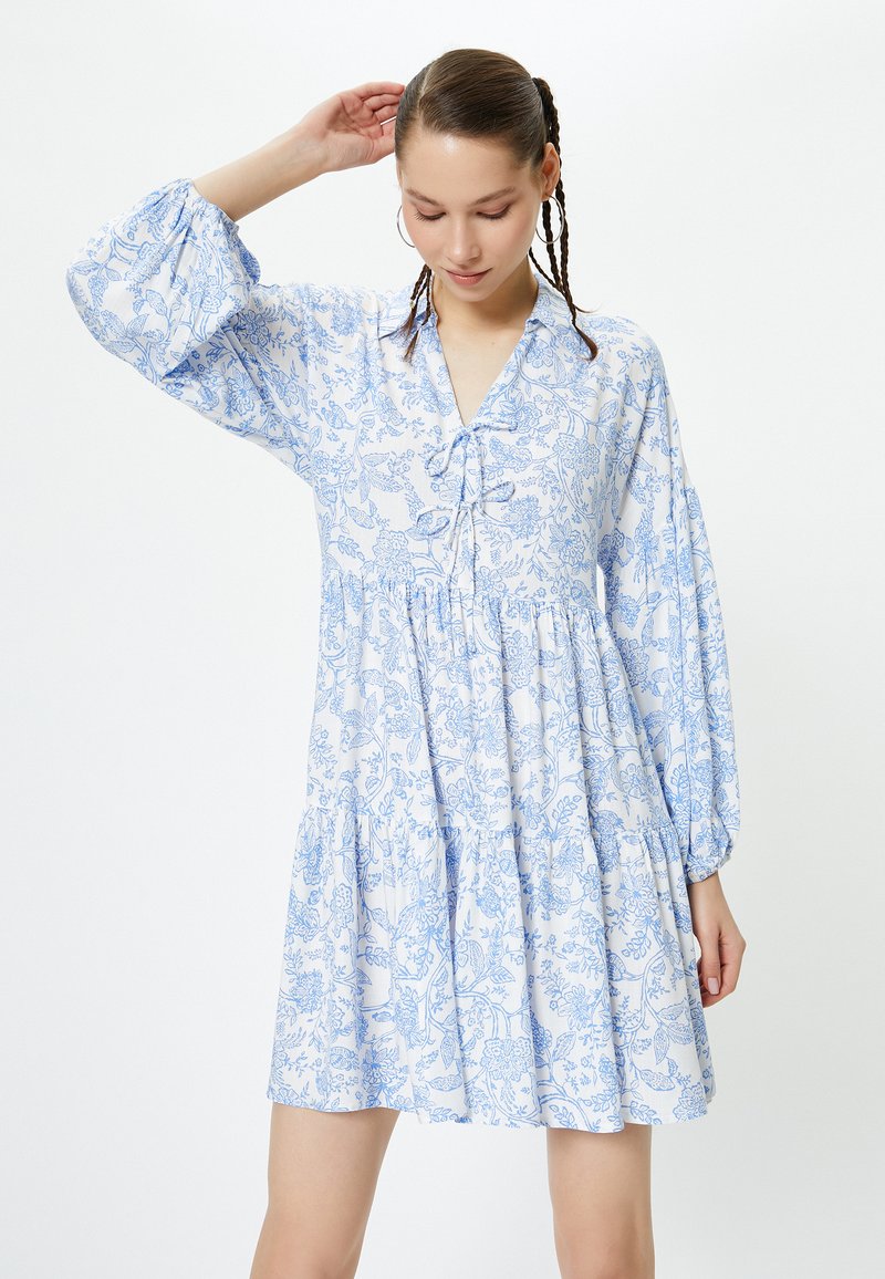 Vestido blanco con estampado floral azul, escote en V y falda fruncida. Las mangas abullonadas añaden volumen. Tela ligera visible en los pliegues.