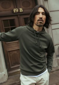 Maslinasto zelena pulover majica s kapuljačom na vezanje, dugim rukavima, mekom teksturom i opuštenim krojem, nošena preko bijele košulje i svijetlih hlača.