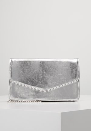 Glanzend zilveren rechthoekige clutch met envelopklep en gedeeltelijk zichtbare kettingriem, geplaatst op een wit oppervlak tegen een effen achtergrond.