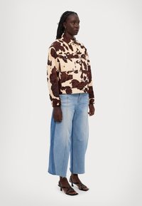 Avskuren jacka med ko-mönster i beige och bruna fläckar, i kombination med ljusblå vida jeans och svarta sandaler med remmar.
