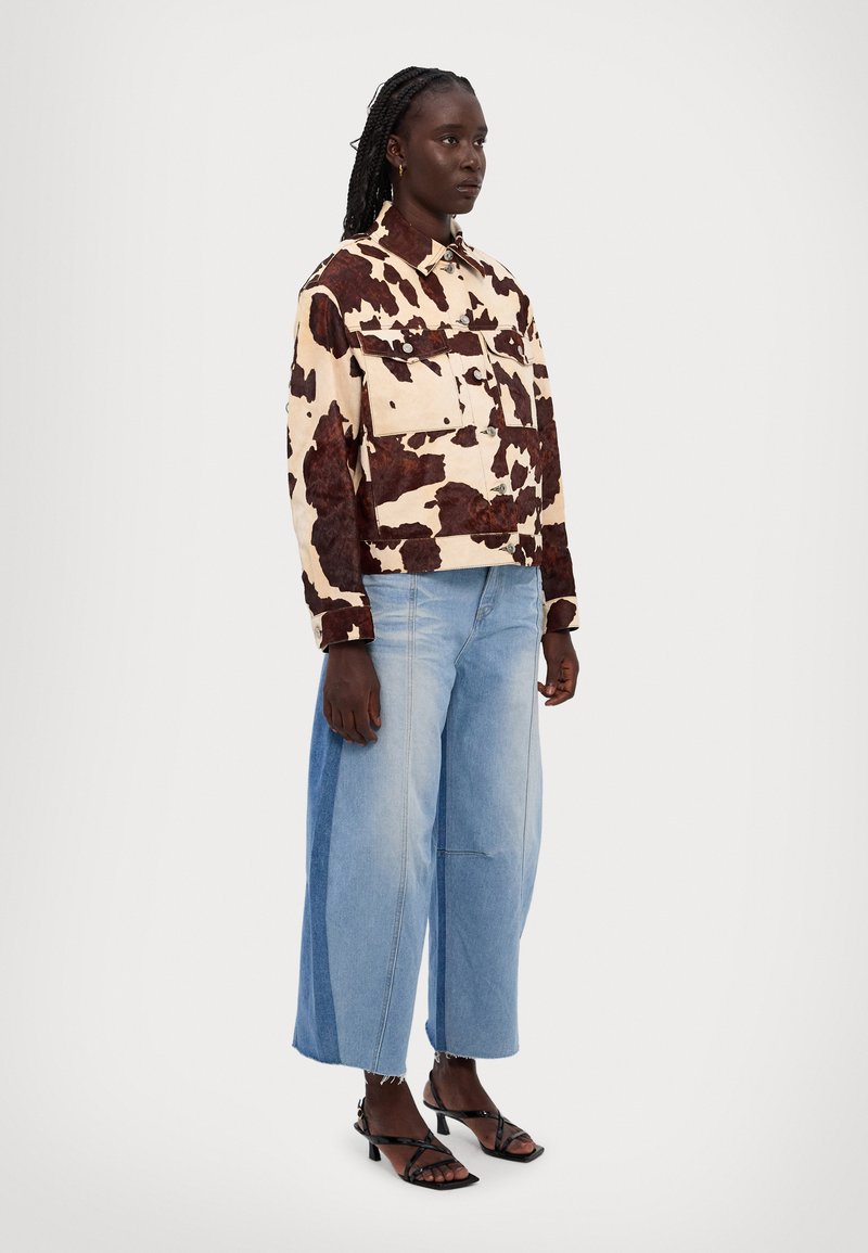 Avskuren jacka med ko-mönster i beige och bruna fläckar, i kombination med ljusblå vida jeans och svarta sandaler med remmar.