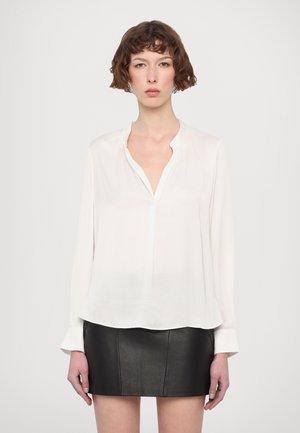 Woman wearing a loose white blouse with a split collar and long sleeves paired with a black leather mini skirt, standing against a plain background.