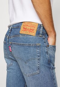 Jeans de mezclilla azul con una etiqueta de cuero en la parte trasera con detalles de la marca y un pequeño logo rojo. Diseño clásico de cinco bolsillos.