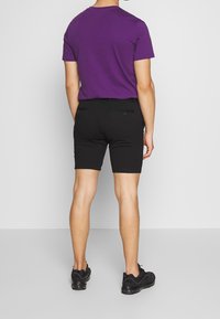 Pantaloni corti neri con una vestibilità su misura e due tasche posteriori, abbinati a una maglietta a maniche corte viola. Modello che indossa sneakers nere.