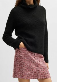 Kvinna iklädd en svart polotröja med långa ärmar och en röd texturerad minikjol, med en hand i fickan, mot en neutral bakgrund.