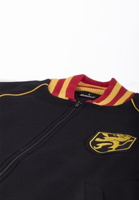 Zwarte ritsjack met een kraag in rood en geel, voorzien van een gele leeuwenom op een schildpatch. Zachte textuur en sportief ontwerp.