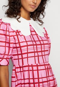 Femme portant une robe à carreaux rose et rouge avec des manches bouffantes et un large col en dentelle blanche, se tenant devant un fond uni.