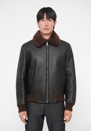 Giacca bomber in pelle nera con colletto in pelo marrone, polsini a coste, chiusura con zip e due tasche laterali. Texture liscia in tutto il capo.