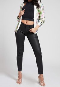 Kvinde iført en blomsterjakke over en sort glitrende crop top, sorte skinny bukser og hvide højhælede sandaler, stående foran en ensfarvet baggrund.