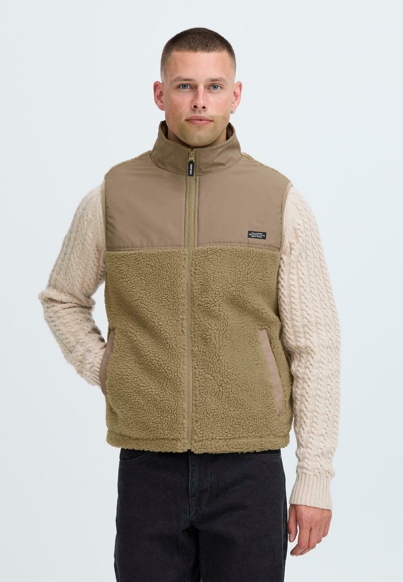 Tan fleece vest with a zip front, high collar, and braided cream sleeves. Features side pockets and a small brand label on chest.