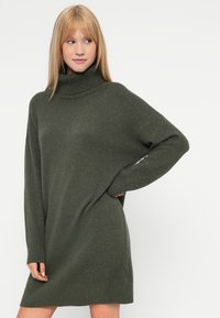 Prevelik pulover obleka s turtleneck ovratnikom v temno olivno zeleni barvi, z rebrastim robom in manšetami, pleteno teksturo ter sproščenim krojem za udobje.