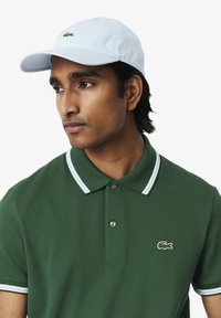 Maglietta polo verde con colletto a righe bianche e blu, con un piccolo logo di coccodrillo ricamato. Cappellino azzurro indossato al contrario.