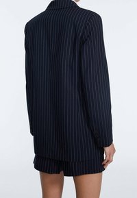 Laivastonsininen liituraitainen bleiseri, joka on valmistettu sileäpintaisesta kankaasta, jossa on kapeakaulus, pitkät hihat ja napilliset kalvosimet. Alla näkyvät shortsit.