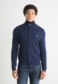 Marinblå zip-up-tröja med hög krage, tillverkad av mjukt tyg, med en liten logotyp på vänster bröst och ribbade detaljer vid ärmsluten.