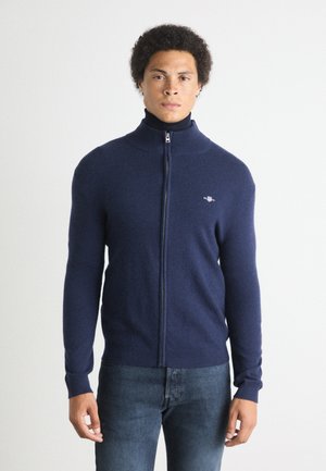 Navy blue zip-up sweater with a high collar, made from soft fabric, featuring a small logo on the left chest and ribbed cuff details.