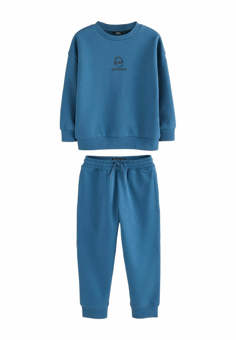 Blå sweatshirt og matchende joggingbukser lavet af blødt materiale. Sweatshirten har en rund hals og et logo-design; joggingbukserne har en elastisk talje.