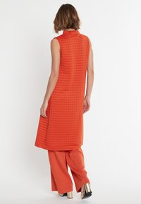 Oranges, gestepptes, ärmelloses Kleid mit hohem Kragen, kombiniert mit passenden, weiten Hosen und metallic glänzenden, hohen Schuhen. Einfaches, strukturiertes Design.