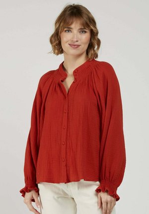 Femme aux cheveux courts et ondulés portant un chemisier rouge ample à manches longues avec des boutons et des poignets à volants, associé à un pantalon de couleur claire.