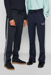 Pantalons de sport bleus marine avec des rayures blanches sur les côtés et une taille élastique, ornés d'un logo sur une jambe. Associés à des baskets foncées.
