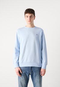 Jeune homme aux cheveux courts et bouclés portant un sweat-shirt bleu clair et un jean bleu sur un fond blanc uni.
