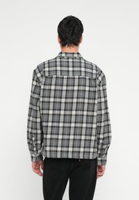 Mies, joka seisoo selin päin ja jolla on musta ja harmaa ruutupaitainen pitkähiha paita sekä mustat housut, valkoista yksiväristä taustaa vasten.