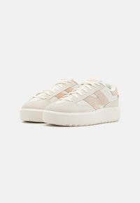 Un par de zapatillas de ante beige y blanco con detalles de ante beige y suelas gruesas blancas sobre un fondo blanco.