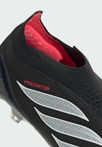 Μαύρο και κόκκινο ποδοσφαιρικό παπούτσι χωρίς κορδόνια με τη σήμανση "Predator" και ασημένιο υφή στο πλάι, που δείχνει τον αστράγαλο και τα καρφιά της σόλας.