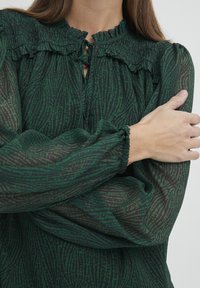 Zelena bluzica s uzorkom s volanom na ovratniku, dugim rukavima i smock detaljima; poluprozirna tkanina s teksturiranim valovitim uzorkom.