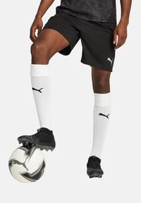 Schwarze Fußballshorts, weiße Kniestrümpfe mit schwarzem Logo und schwarze Fußballschuhe, die auf einem schwarz-weißen gemusterten Fußball stehen.