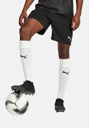 Black football shorts, white knee-high socks with black logo, and black football boots, standing on a black and white patterned football.