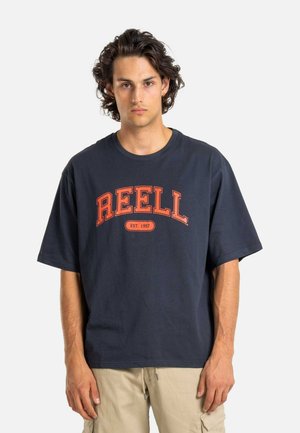 Joven con cabello rizado oscuro, vistiendo una camiseta de gran tamaño en color azul marino con "REELL EST. 1997" en naranja, y pantalones cortos de carga beige, frente a un fondo blanco.