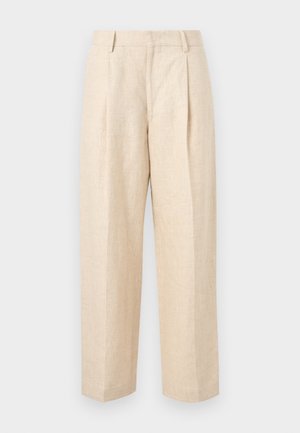 Homme debout en pantalon beige à jambes larges, chemise blanche rayée à manches longues retroussées, et chaussures noires, mains dans les poches, regardant vers la gauche.