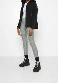 Black softshell jacket with hood, white top, light green melange leggings, and black hiking boots with silver accents and laces.