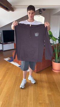 Giovane uomo che tiene un felpa scura con una grafica di un sole sorridente in un soggiorno con pavimento in legno, divano rosso e pianta verde in vaso.