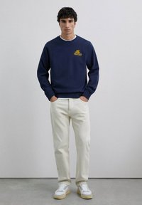 Merimetsäntyylinen collegepaita, jossa on keltainen logo, puuvillakangasta, pyöreä kaula-auko ja resorit hihansuissa, yhdistettynä kermaisiin housuihin ja valkoisiin tennareihin.