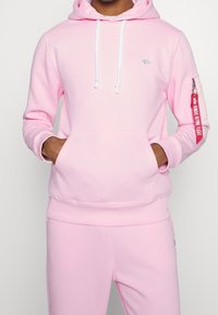 Man som bär en ljusrosa hoodie och matchande sweatpants, med händerna i hoodiefickan, vita dragsnören och en röd "Remove Before Flight"-etikett på ärmen.