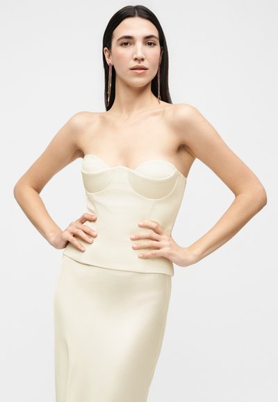 Femme aux longs cheveux foncés porte un haut corset crème sans bretelles et une jupe assortie, les mains posées sur les hanches, avec de longues boucles d’oreilles sur fond uni.