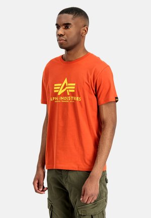 Uomo che indossa una t-shirt arancione Alpha Industries e pantaloni cargo verdi in piedi davanti a uno sfondo semplice.