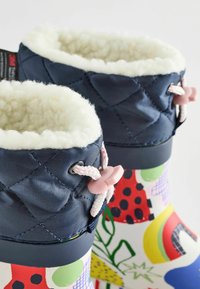 Bottes de pluie colorées avec un haut matelassé bleu marine, doublure en fausse fourrure, détails roses et un bas en caoutchouc à motifs présentant différentes formes et couleurs.