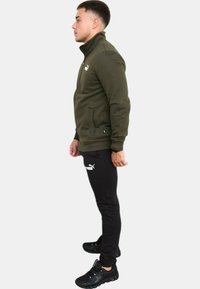 Giacca verde con zip e colletto alto, abbinata a pantaloni neri elasticizzati con logo bianco e scarpe da ginnastica nere.