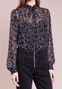 Femme portant un chemisier noir transparent à motifs d'étoiles blanches et un col lavallière, associé à un pantalon noir taille haute avec une fermeture éclair à l'avant.