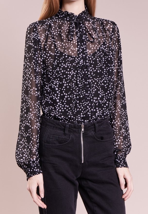 Femme portant un chemisier noir transparent à motifs d'étoiles blanches et un col lavallière, associé à un pantalon noir taille haute avec une fermeture éclair à l'avant.