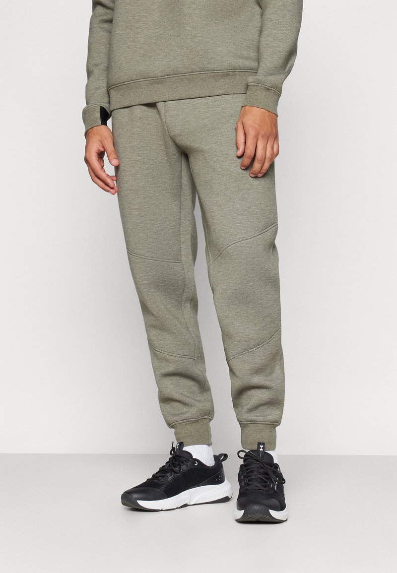 Maslinasto zeleni dukserica s okruglim izrezom u kombinaciji s odgovarajućim suknjim hlačama koje imaju rebraste manžete i teksturirane panele. Crne atletske cipele.