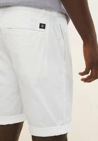 Pantaloni shorts bianchi in cotone con risvolti, dotati di una tasca sul retro e di una piccola etichetta nera con logo. Texture liscia e vestibilità sartoriale.
