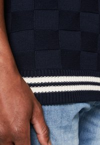 Pull en maille bleu marine avec un motif à carreaux, des poignets côtelés et un ourlet rayé blanc. Main visible en bas, reposant sur du denim.