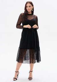 Czarna midi sukienka z przezroczystymi, długimi rękawami, ozdobiona leopardzim wzorem oraz związaną talią. Całość dopełniają szpilki.