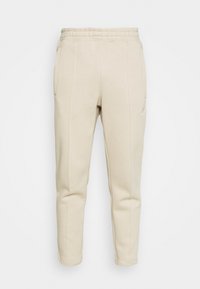 Béžové tapered tepláky s elastickým pasem a bočními kapsami, s jemnými vertikálními švy a malým logem na pravém stehně.