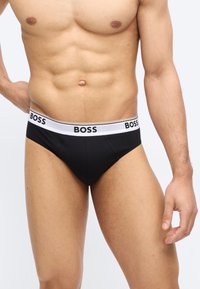 Αντρικός κορμός με μαύρα εσώρουχα BOSS με λευκή ζώνη μέσης, που δείχνει καθορισμένους κοιλιακούς μύες και χαλαρά χέρια, σε απλό φόντο.